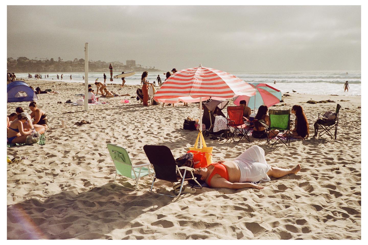 A woman in an orange top and white skirt lies on a sandy beach with other people and umbrellas in the background.