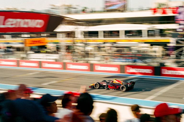 A race car speeds along a track with spectators in the foreground and buildings in the background.