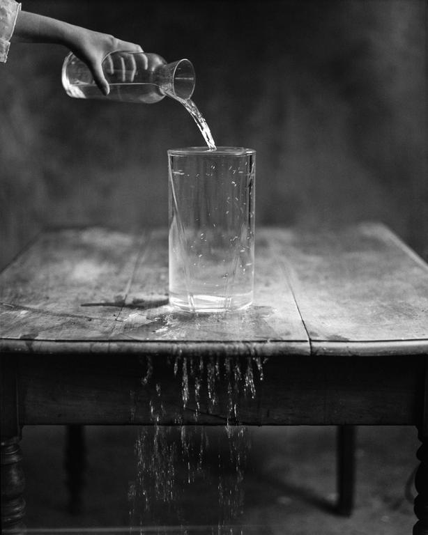 A hand pours water from a carafe into a glass on a wooden table, with water spilling over the side of the table.