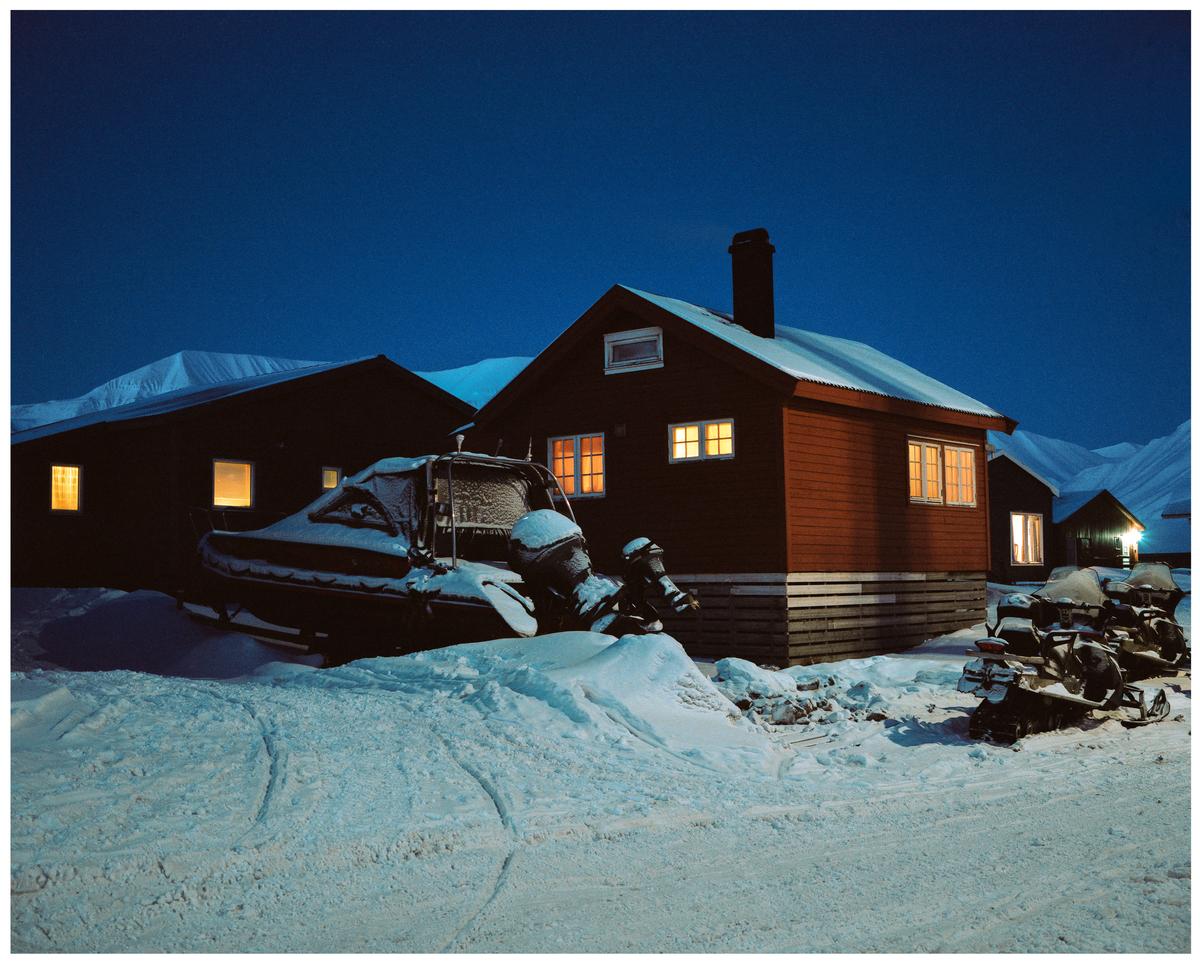 A snow-covered boat and snowmobiles are parked outside houses with illuminated windows in a snowy landscape at night.