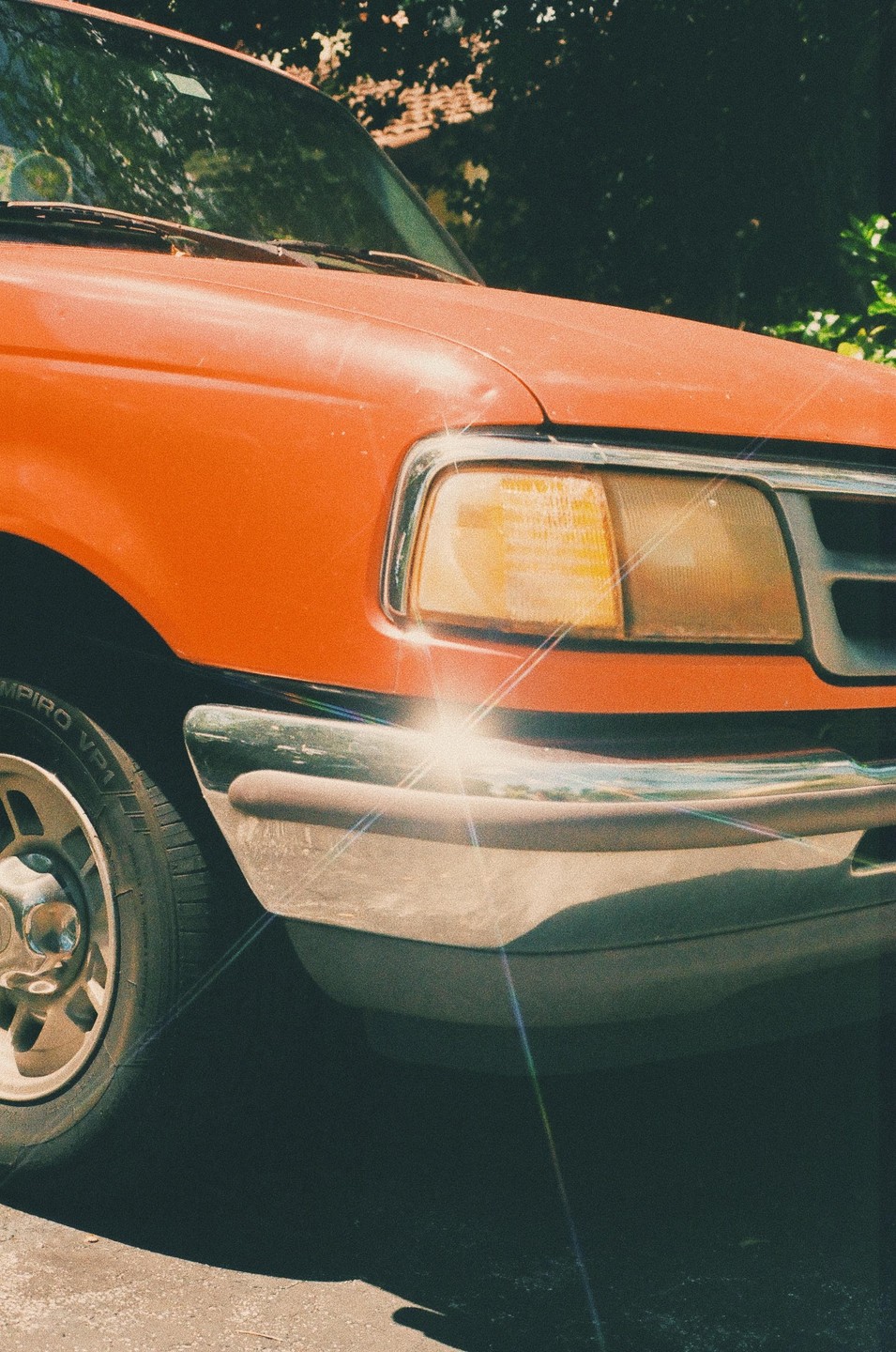 The front of an orange pickup truck is parked outside, with sunlight reflecting off its bumper and headlight.