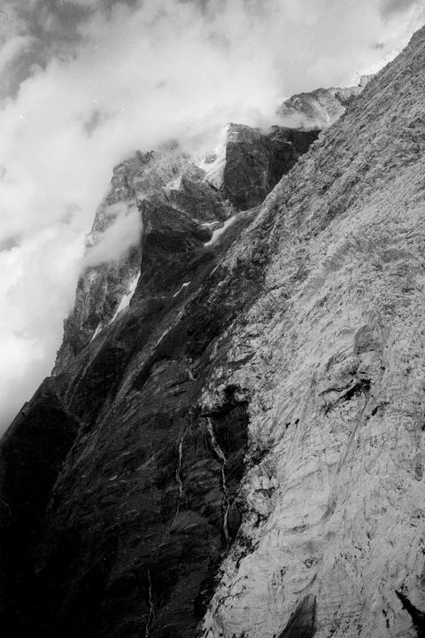 Clouds partially obscure a rugged mountain peak and a glacier in the French Alps.