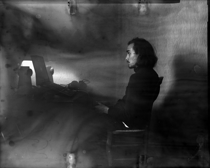 A man with long hair sits on a chair next to a laptop and headphones.