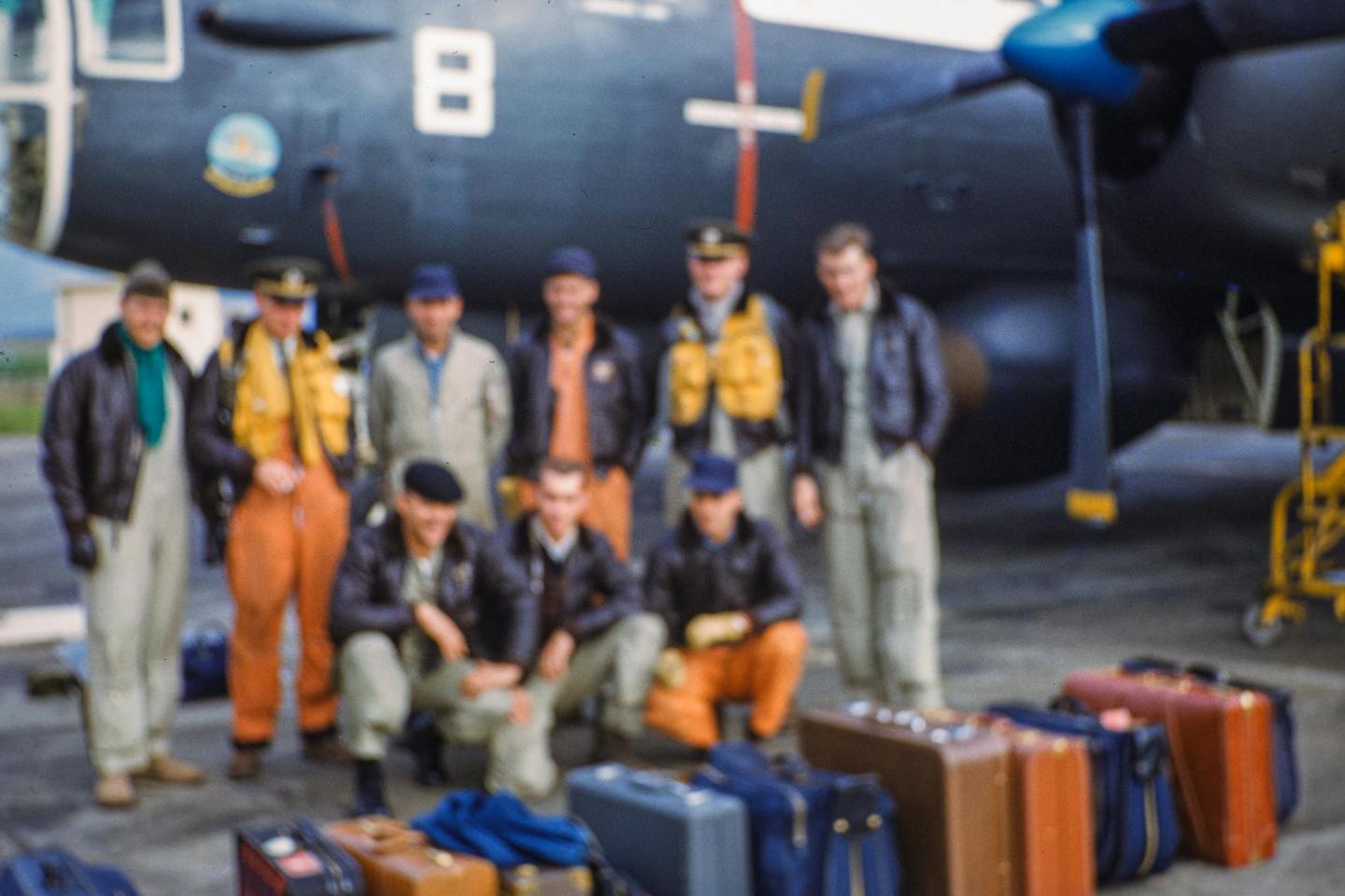 A group of men in uniform stand and crouch in front of an airplane with several suitcases on the ground.