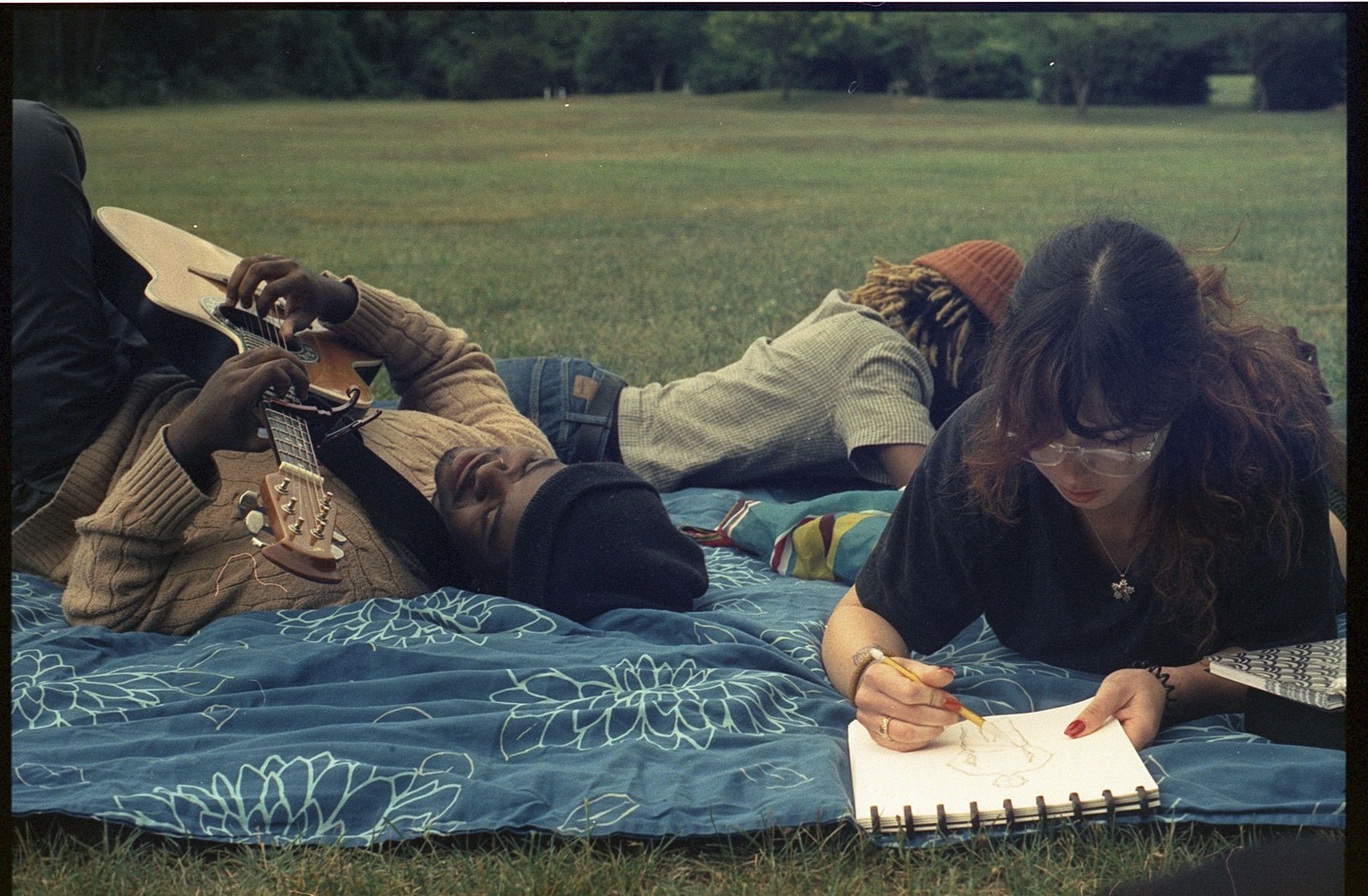 A man plays guitar while lying on a blue blanket in a park, and a woman draws in a sketchbook next to him.