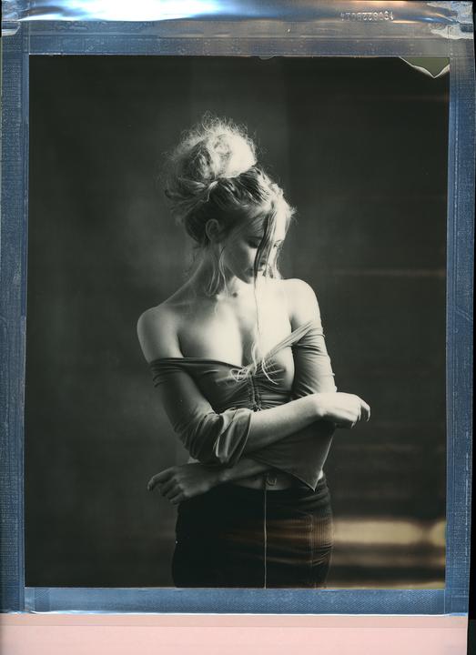 A woman with her hair in a bun and wearing an off-the-shoulder top looks down with her arms crossed.