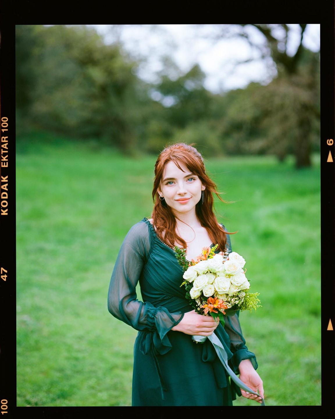 A woman with red hair and a green dress holds a bouquet of white roses in a grassy meadow.