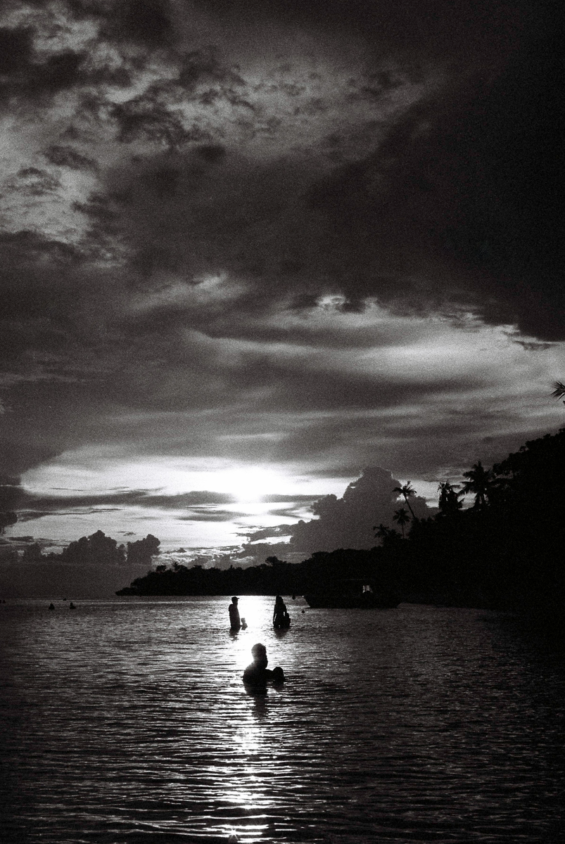 Three people are in the water with a boat and an island with palm trees in the background under a dramatic sky.