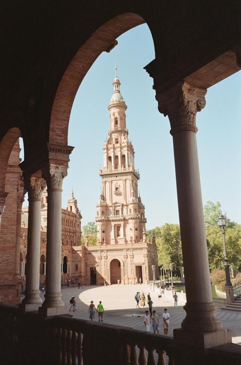 A tall, ornate tower rises above a plaza with people walking around, framed by dark arches and columns in the foreground.