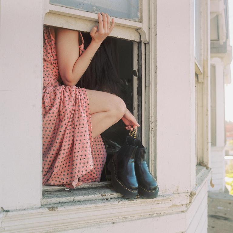 A woman in a pink dress sits in a window frame, holding a pair of black boots.