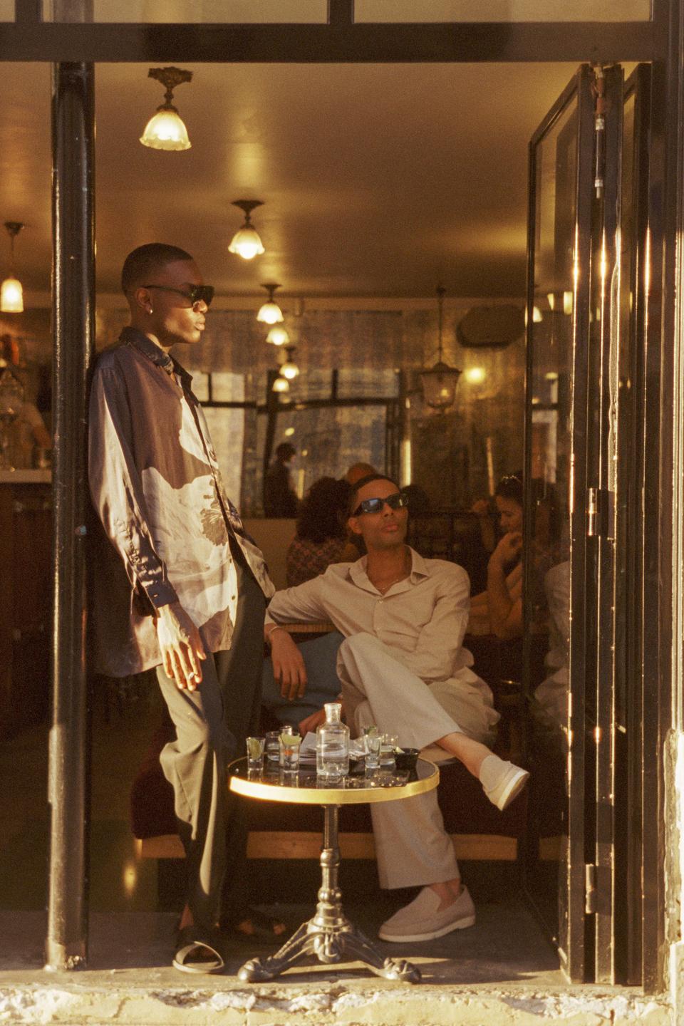 Two men are relaxing at a cafe table with drinks, one standing and one sitting, with other patrons visible in the background.