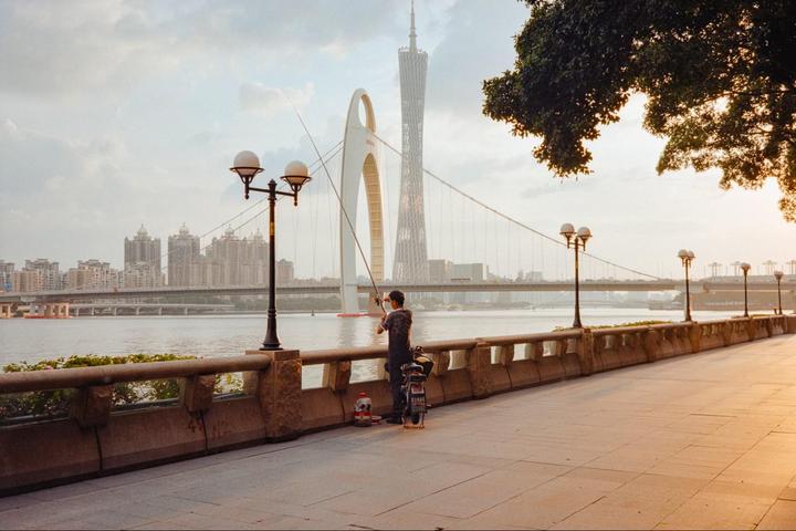 A man fishes from a riverbank promenade with a city skyline and bridge in the background at sunset.