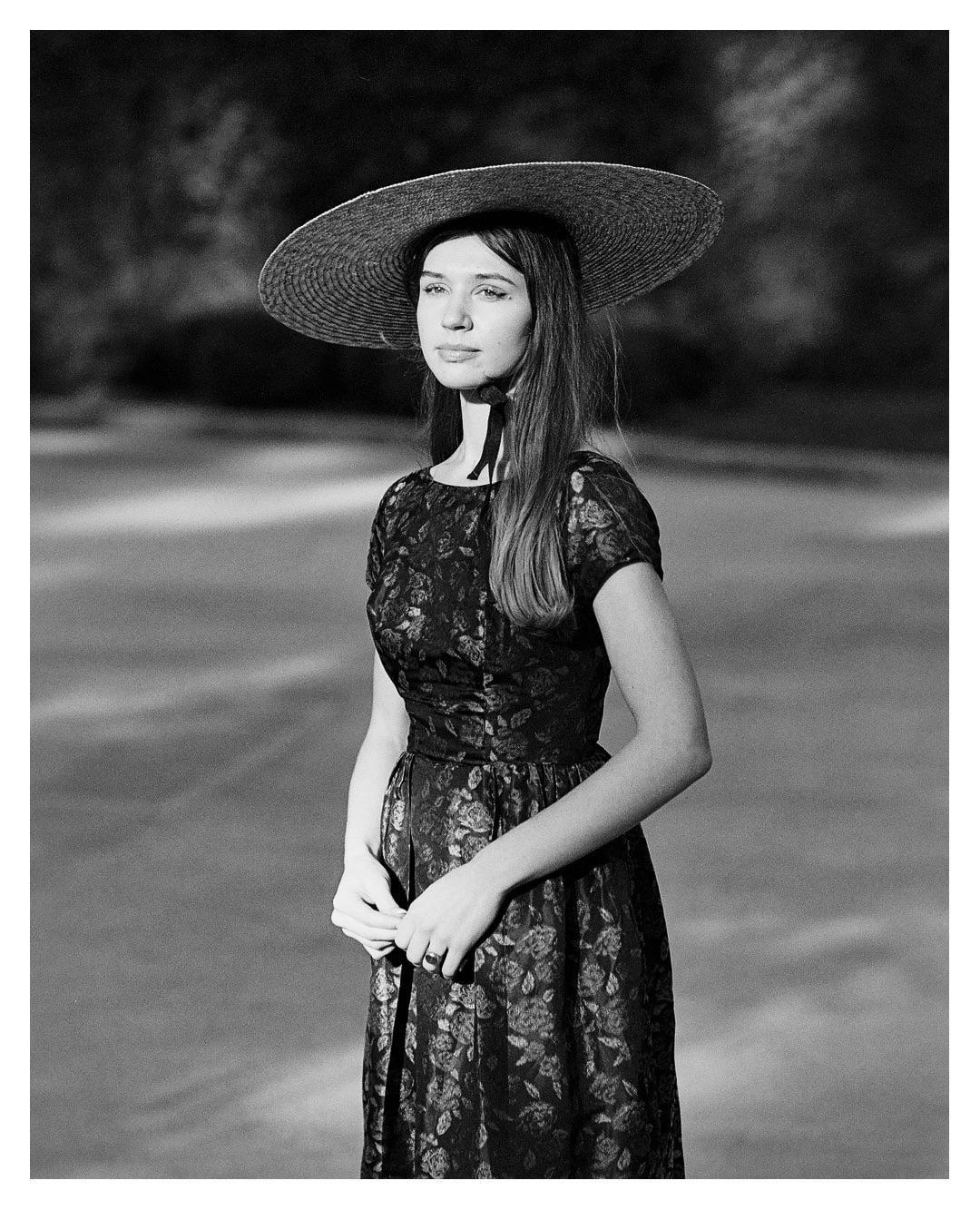 A woman wearing a wide-brimmed hat and a patterned dress stands outdoors.