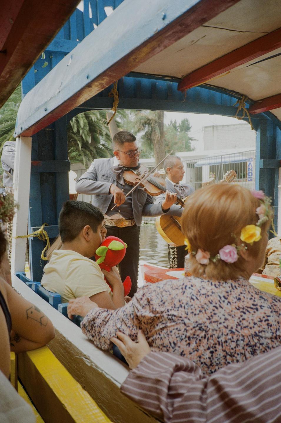 Two musicians play violin and guitar on a boat while a woman with a flower crown and a boy holding a toy listen.