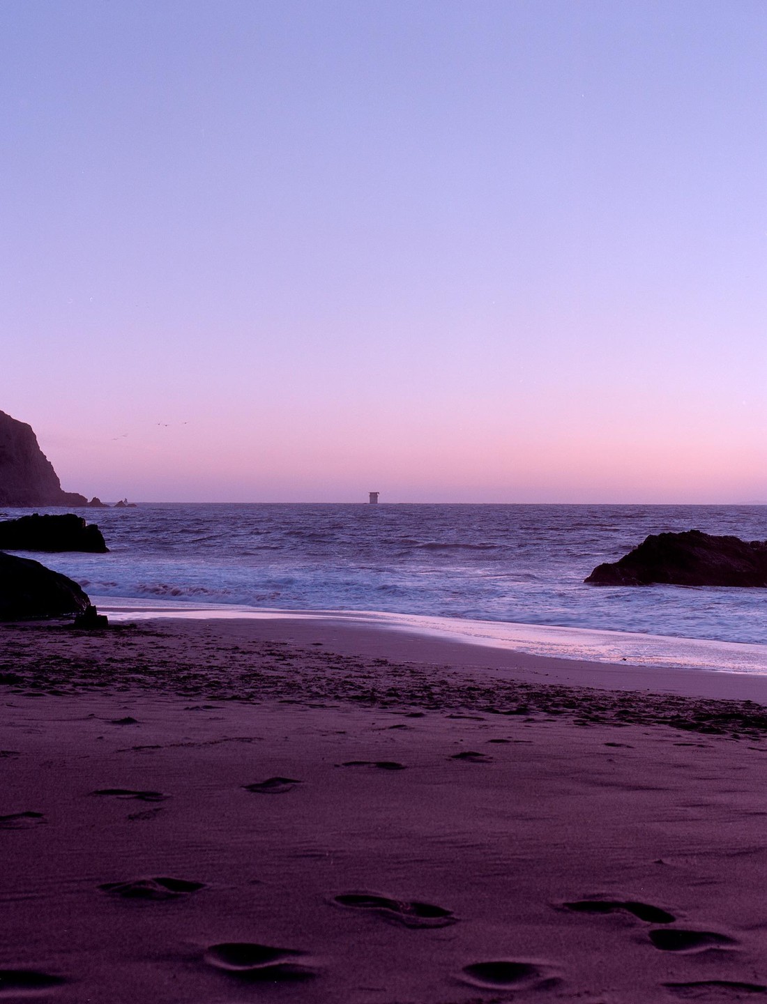 Footprints are visible in the sand on a beach with waves crashing on the shore and a distant structure in the ocean under a purple sky.
