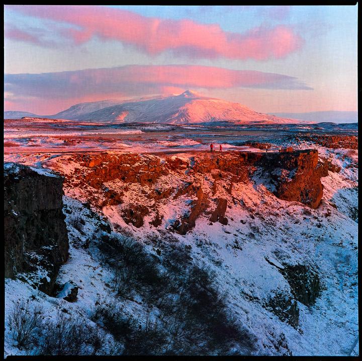Two people stand on a snowy cliff overlooking a vast, mountainous landscape under a sky with pink clouds.