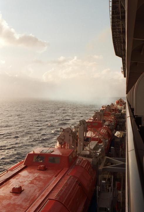 A row of red lifeboats are visible on the side of a large ship, with the ocean and a hazy sky in the background.