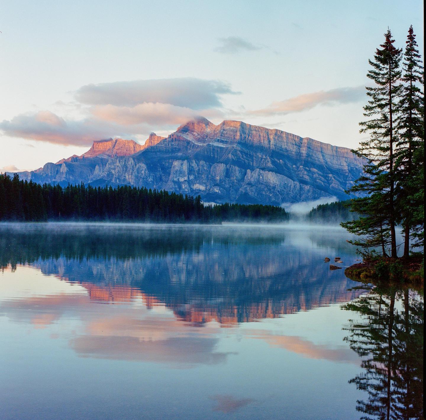 A mountain range with a forest at its base is reflected in a misty lake at dawn.