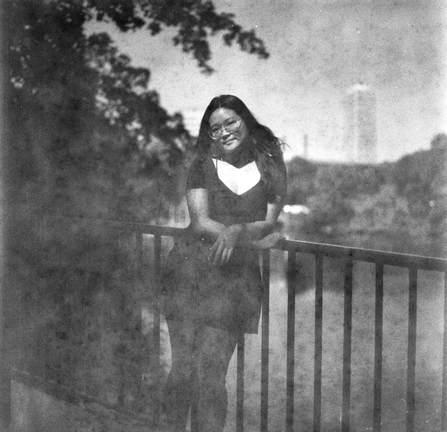 A woman with long hair and glasses leans on a railing with a city skyline in the background.