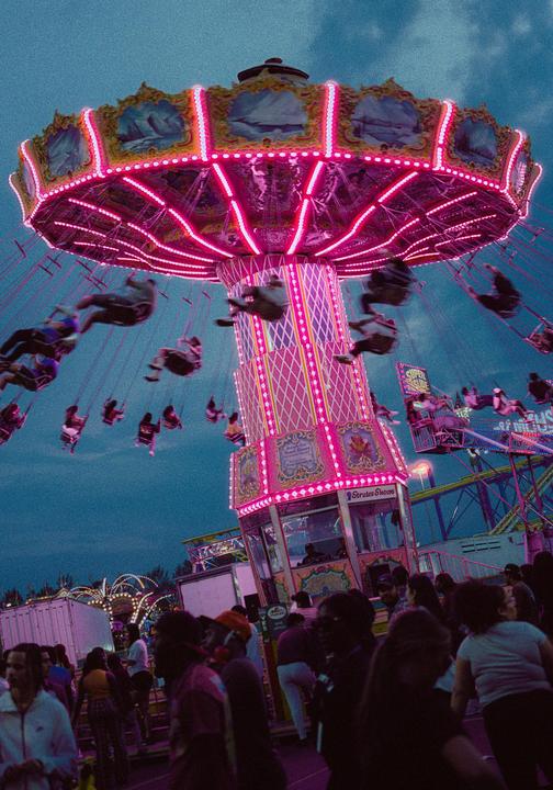 A swing ride with many people is lit up in pink and yellow at a fairground at dusk.