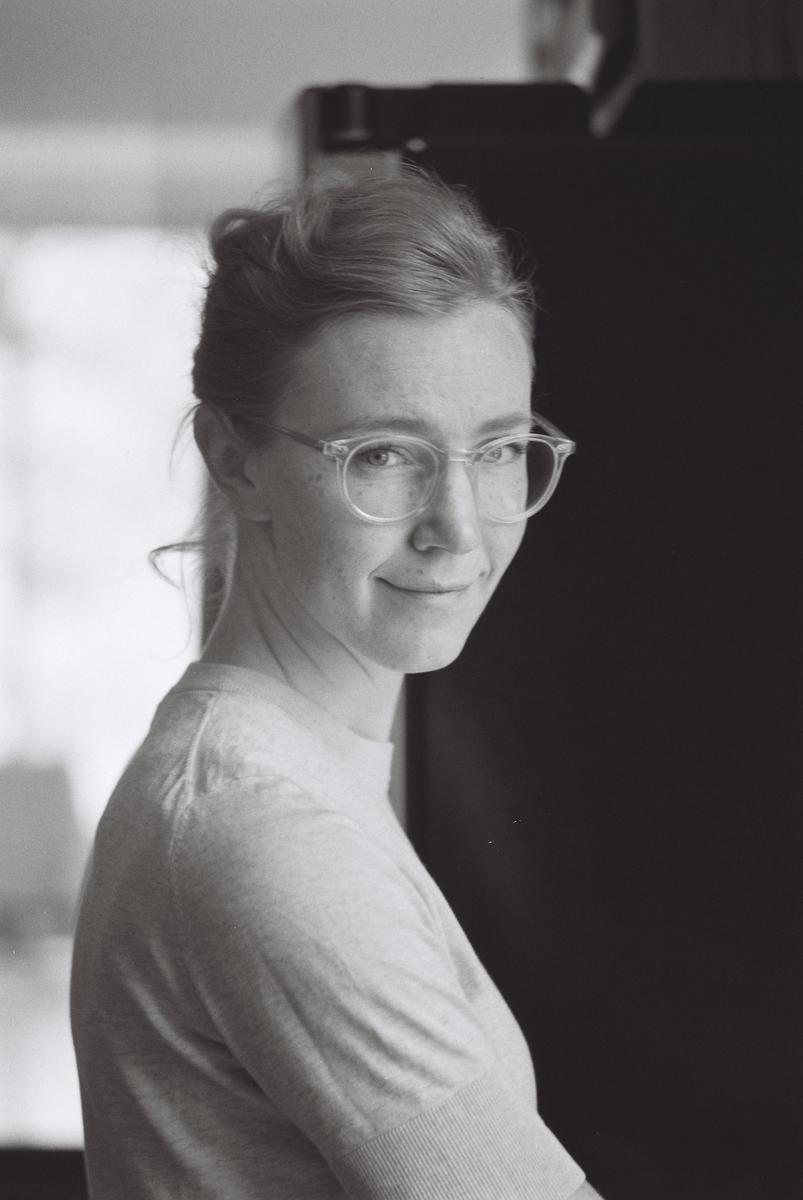 A woman with glasses and her hair tied back smiles at the viewer in a self-portrait.