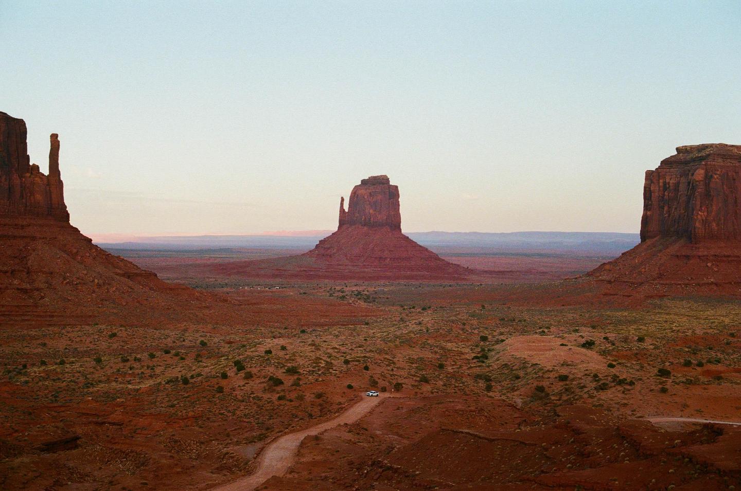 A vast desert landscape features towering rock formations under a pale sky with a dirt road leading towards a distant mesa.