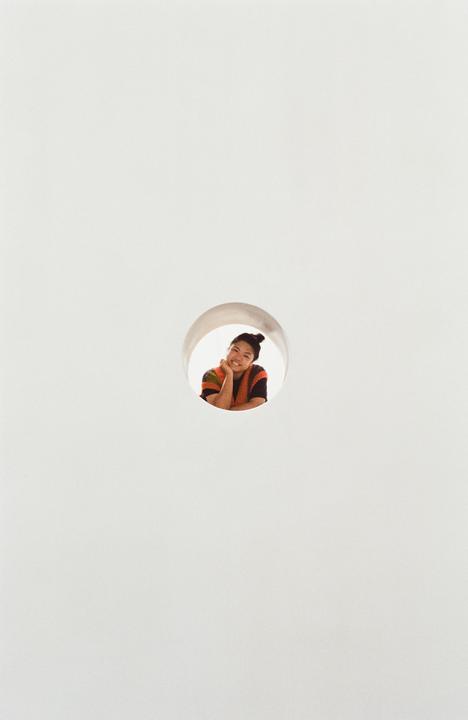 A woman with her hair in a bun smiles while looking through a circular opening in a white wall.