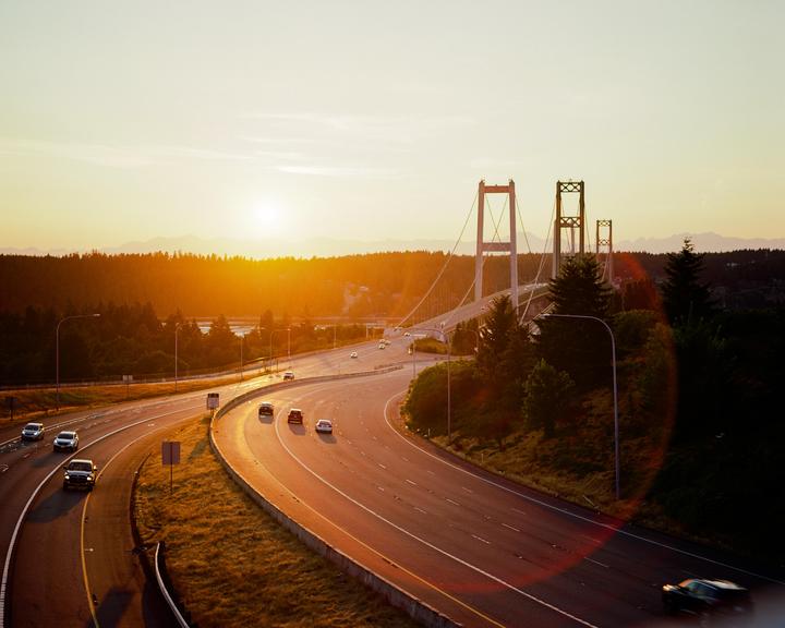 A highway curves towards a suspension bridge at sunset, with cars driving on the road.