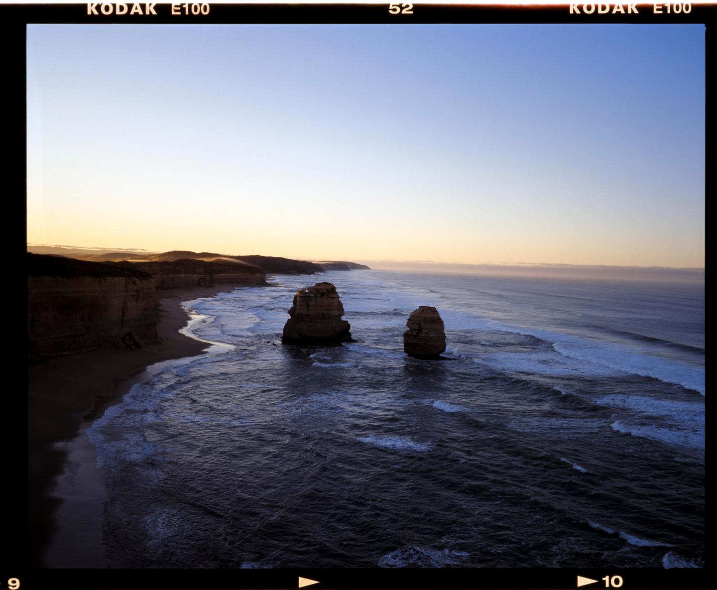 Two large rock formations stand in the ocean near a coastline with cliffs and waves.