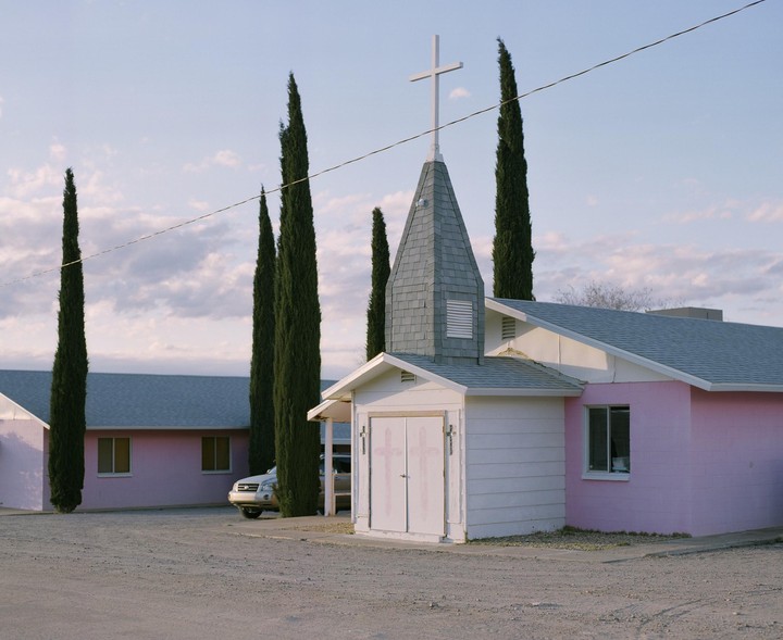 A pink church with a steeple and cross stands next to tall, thin trees under a cloudy sky.