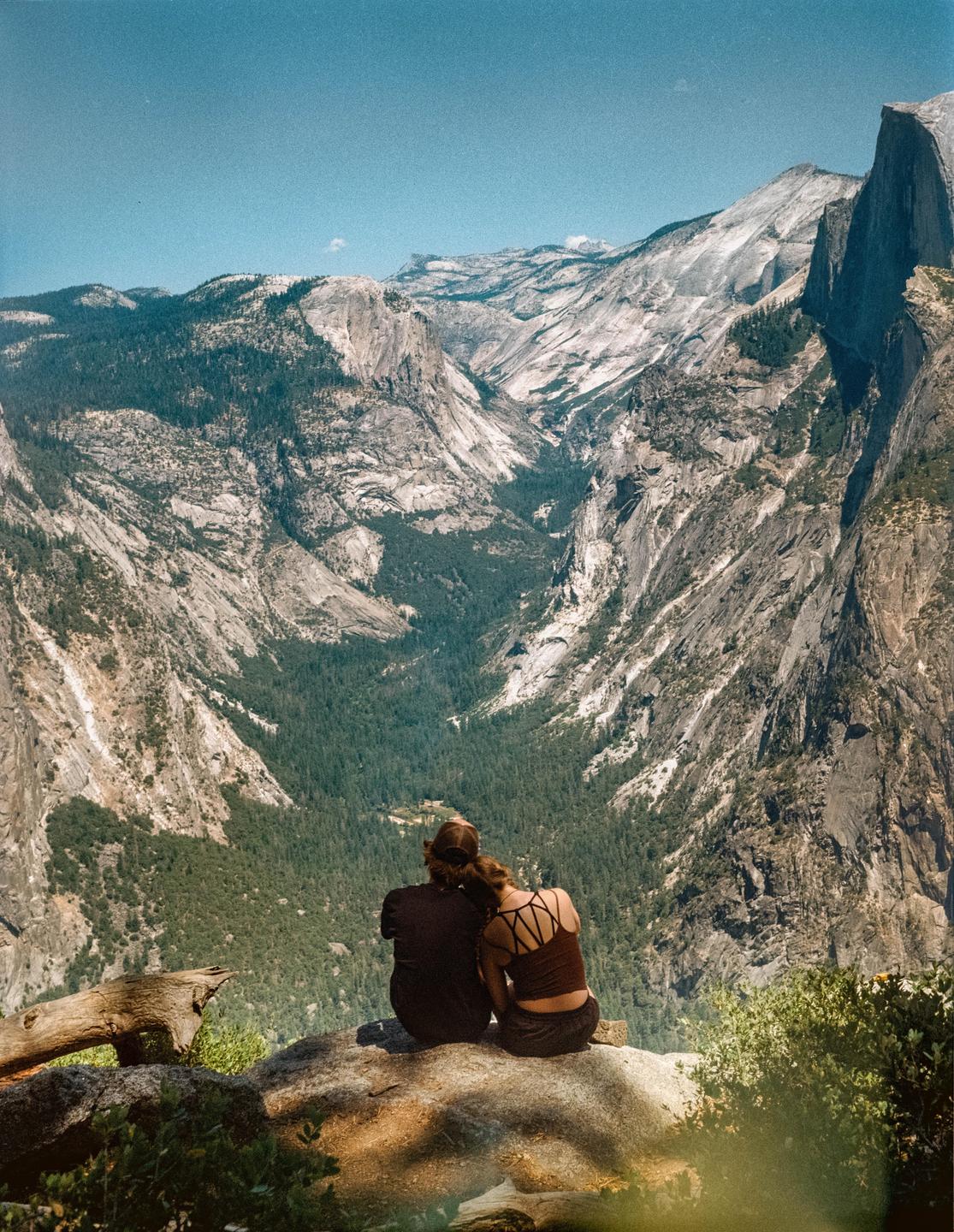 A couple sits on a rock overlooking a vast mountain valley.
