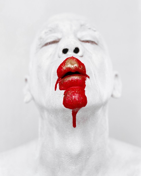 A person with white body paint has red glitter lipstick dripping from their mouth.