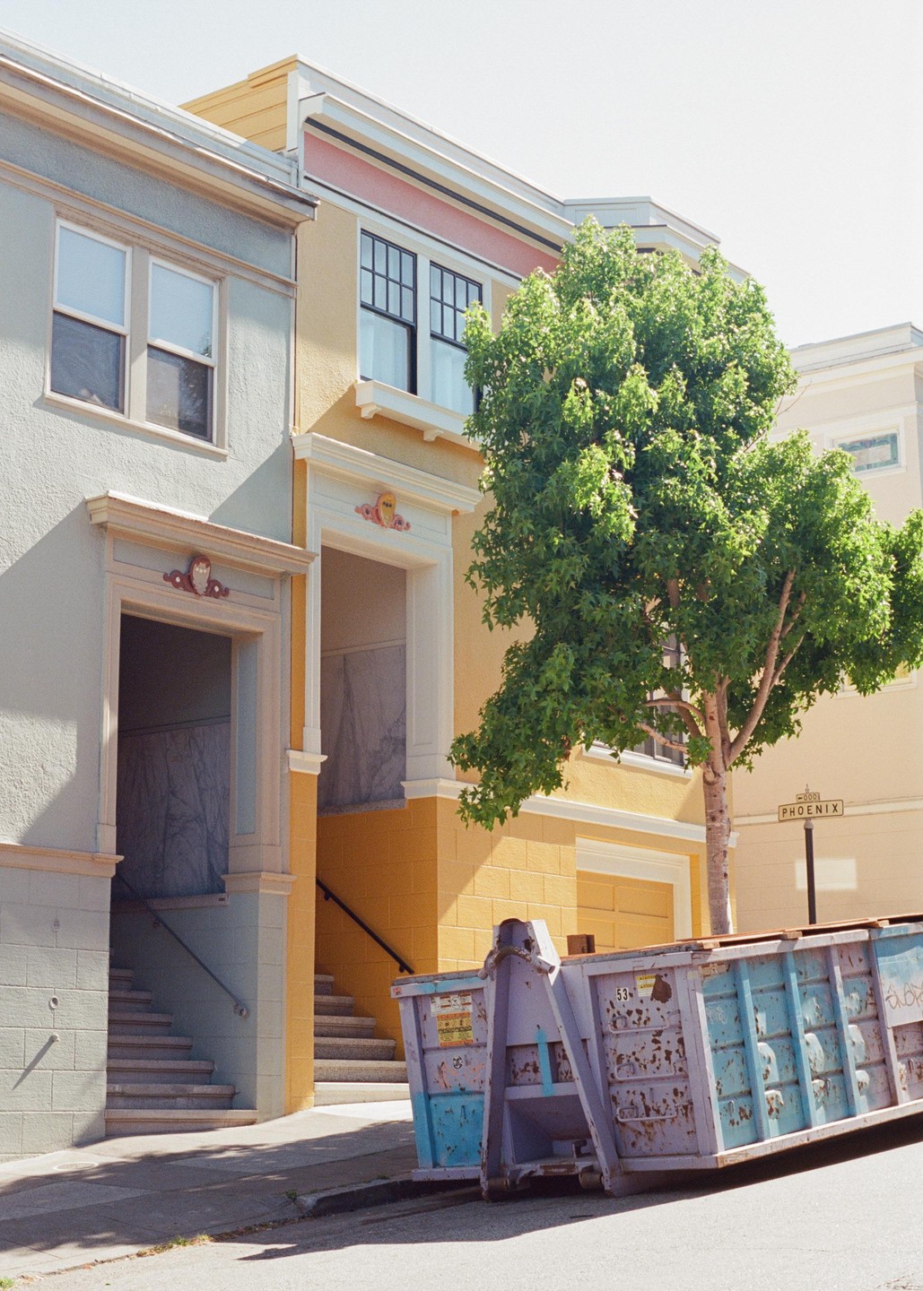 A blue and purple dumpster sits on a street in front of a tree and two buildings with stairs leading to their entrances.