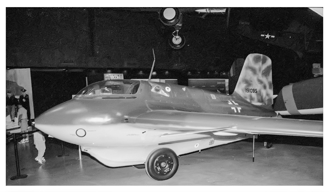 A black and white photograph shows a Messerschmitt Me 163 Komet rocket-powered interceptor aircraft on display in a museum.