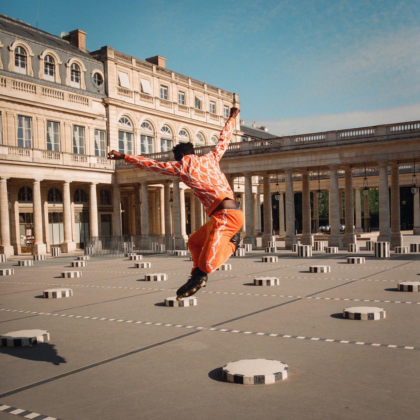 A man in orange clothing and rollerblades jumps in a courtyard with black and white striped pillars and a large building in the background.