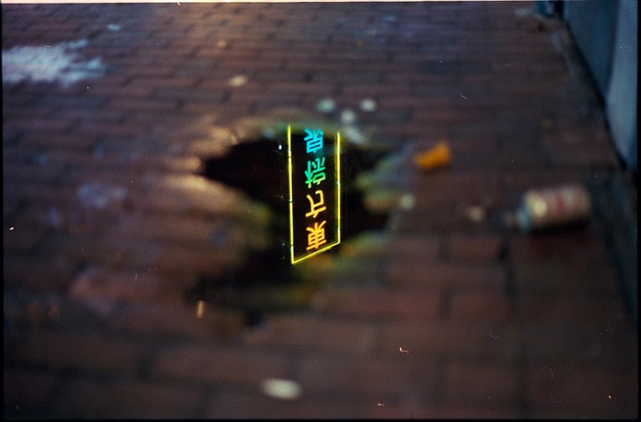 A neon sign with Japanese characters is reflected in a puddle on a brick street at night.