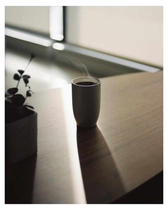 A steaming cup of coffee sits on a wooden table next to a potted plant, with a window in the background.
