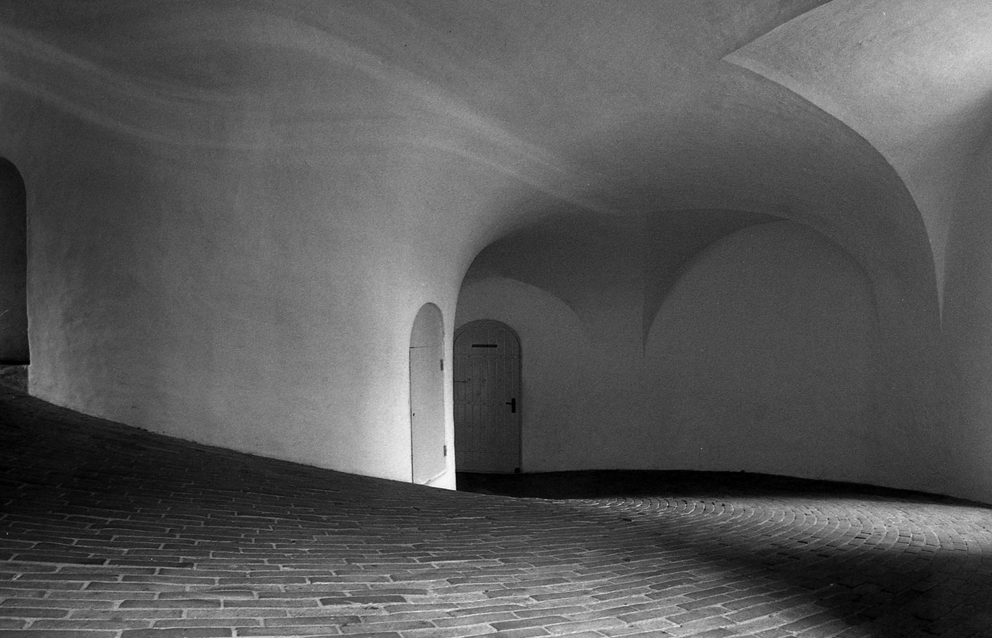 A brick-paved, arched hallway with two doors is illuminated by light from the right.