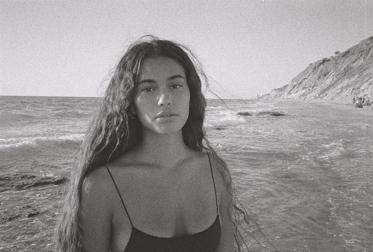 A woman with long, wavy hair stands in the ocean with a cliff and beach in the background.