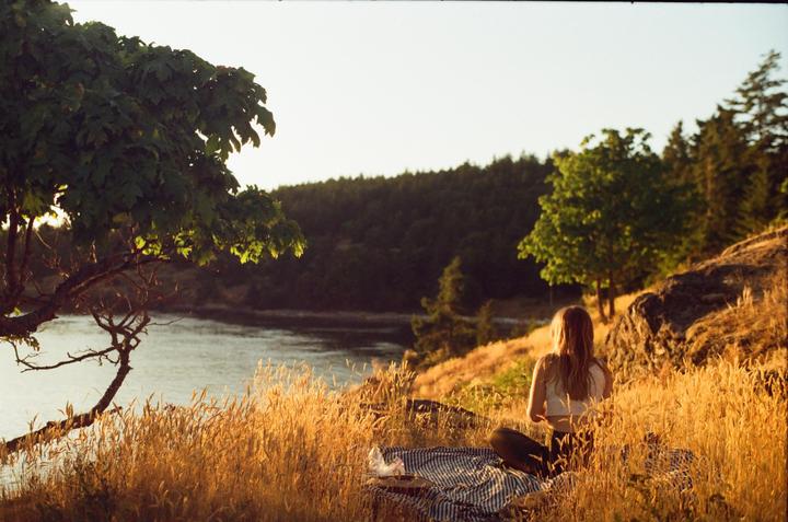A woman sits on a blanket in a field of tall grass overlooking a body of water at sunset.