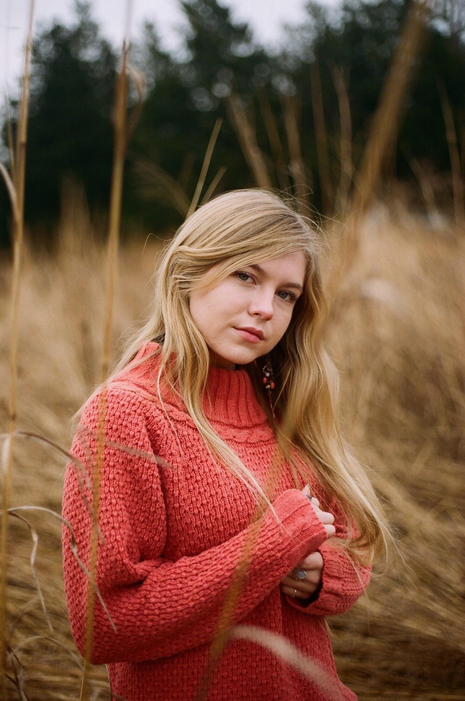 A woman with long blonde hair wears a coral sweater and stands in a field of tall, dry grass.