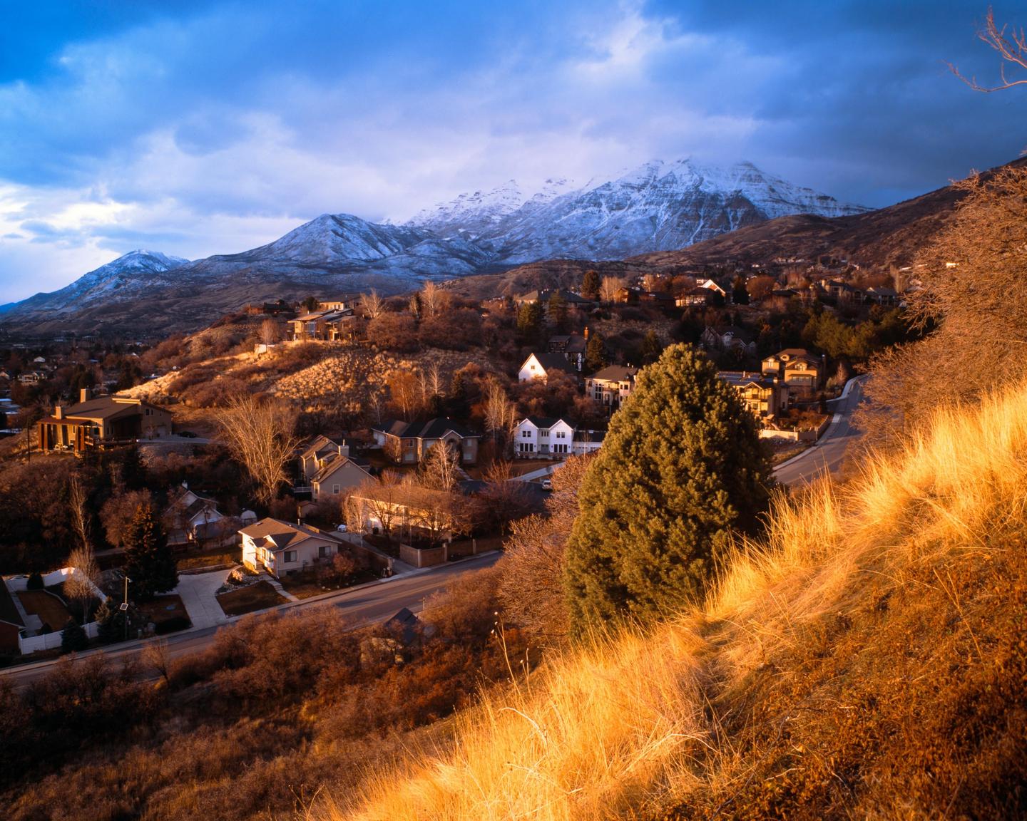 A hillside neighborhood with houses and trees is illuminated by golden light, with snow-capped mountains and a cloudy sky in the background.