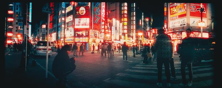 A street in Tokyo is bustling with people and illuminated by numerous neon signs at night.