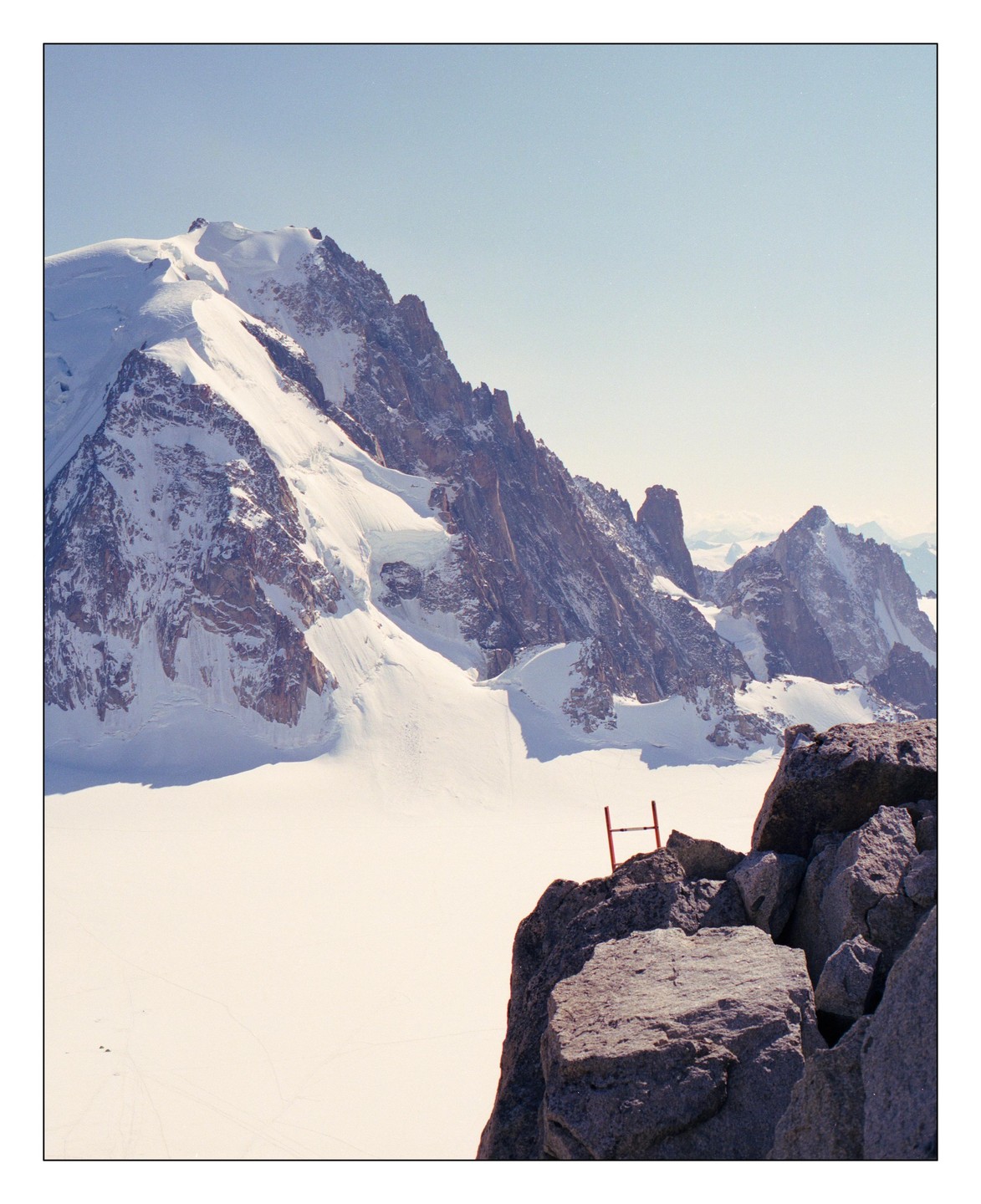 A vast snowfield stretches out before a rugged mountain range, with a small red structure visible on a rocky outcrop in the foreground.