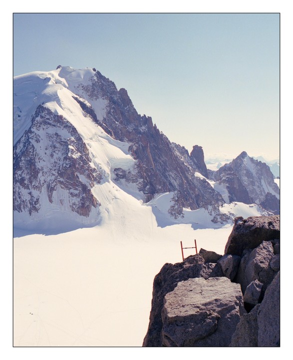 A vast snowfield stretches out before a rugged mountain range, with a small red structure visible on a rocky outcrop in the foreground.