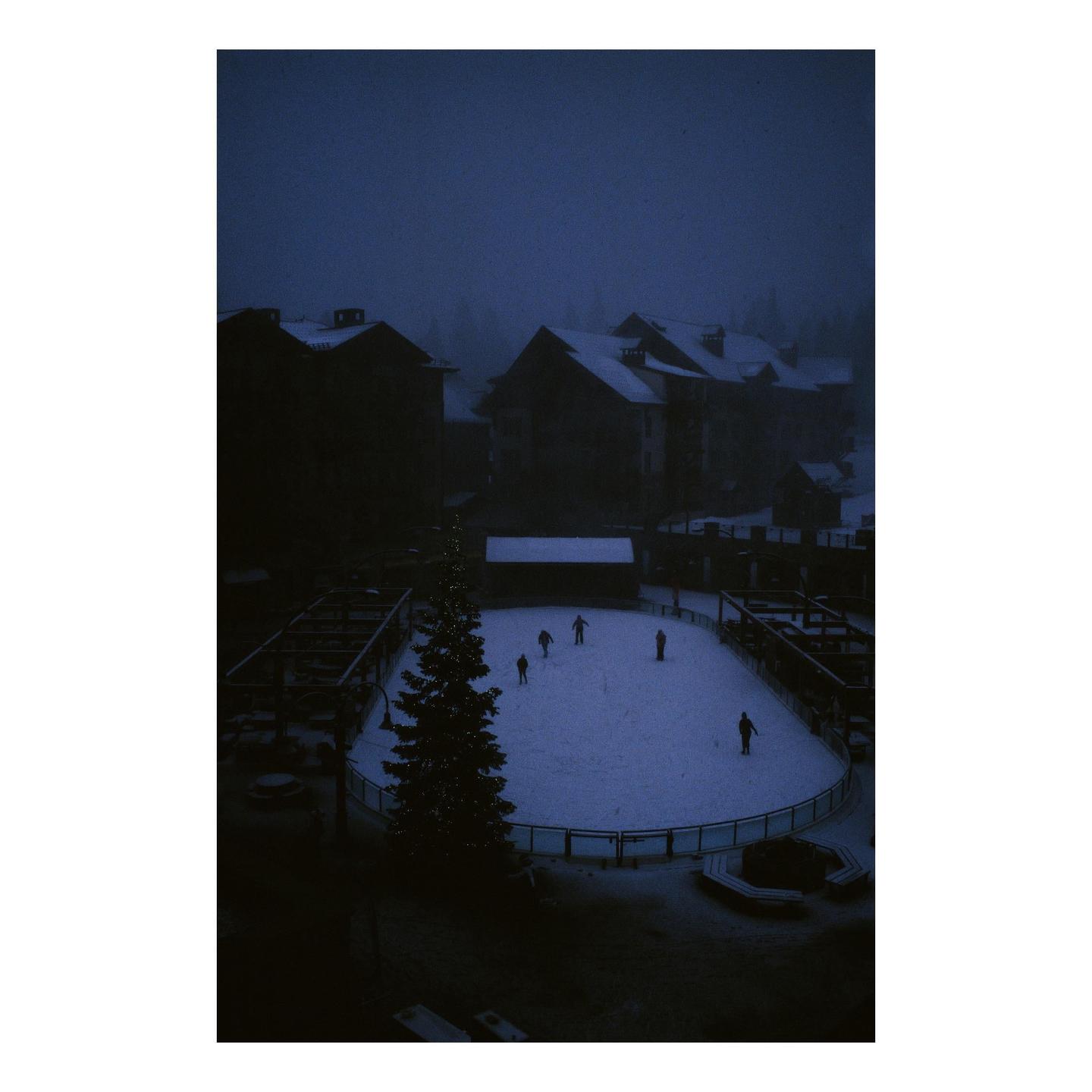 A group of people are ice skating on an outdoor rink next to a large Christmas tree at night.