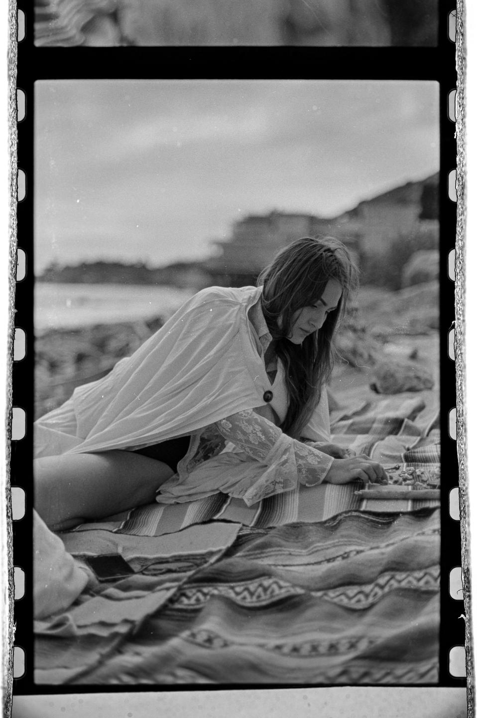A woman in a white shirt and dark bottoms is lying on a patterned blanket on a rocky beach.
