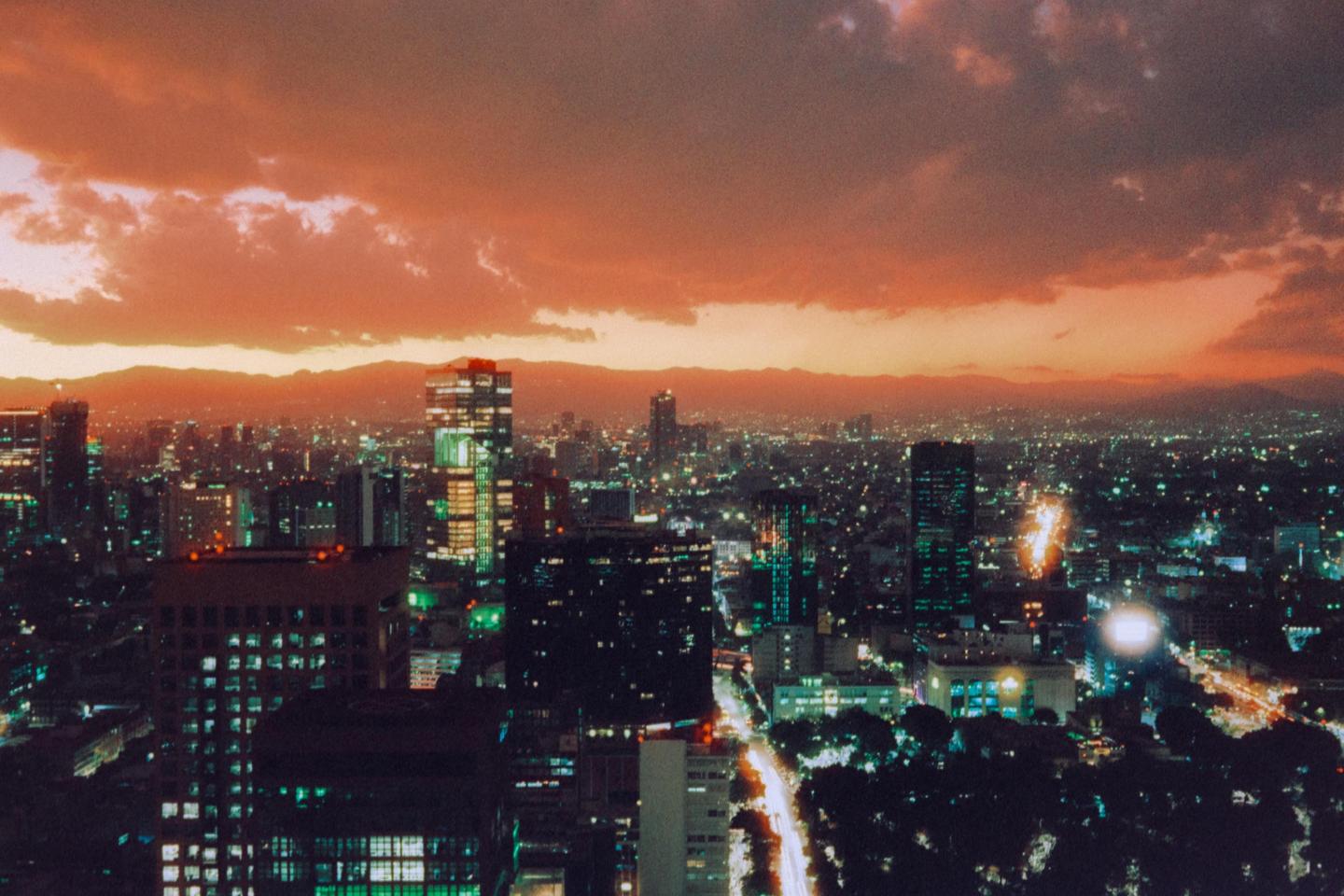 A panoramic view of Mexico City shows a cityscape with illuminated buildings under a dramatic orange and purple sky at dusk.