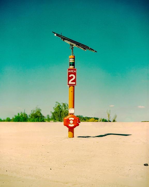 A yellow pole with a solar panel and an "AREA 2" sign stands on a sandy beach under a clear blue sky.