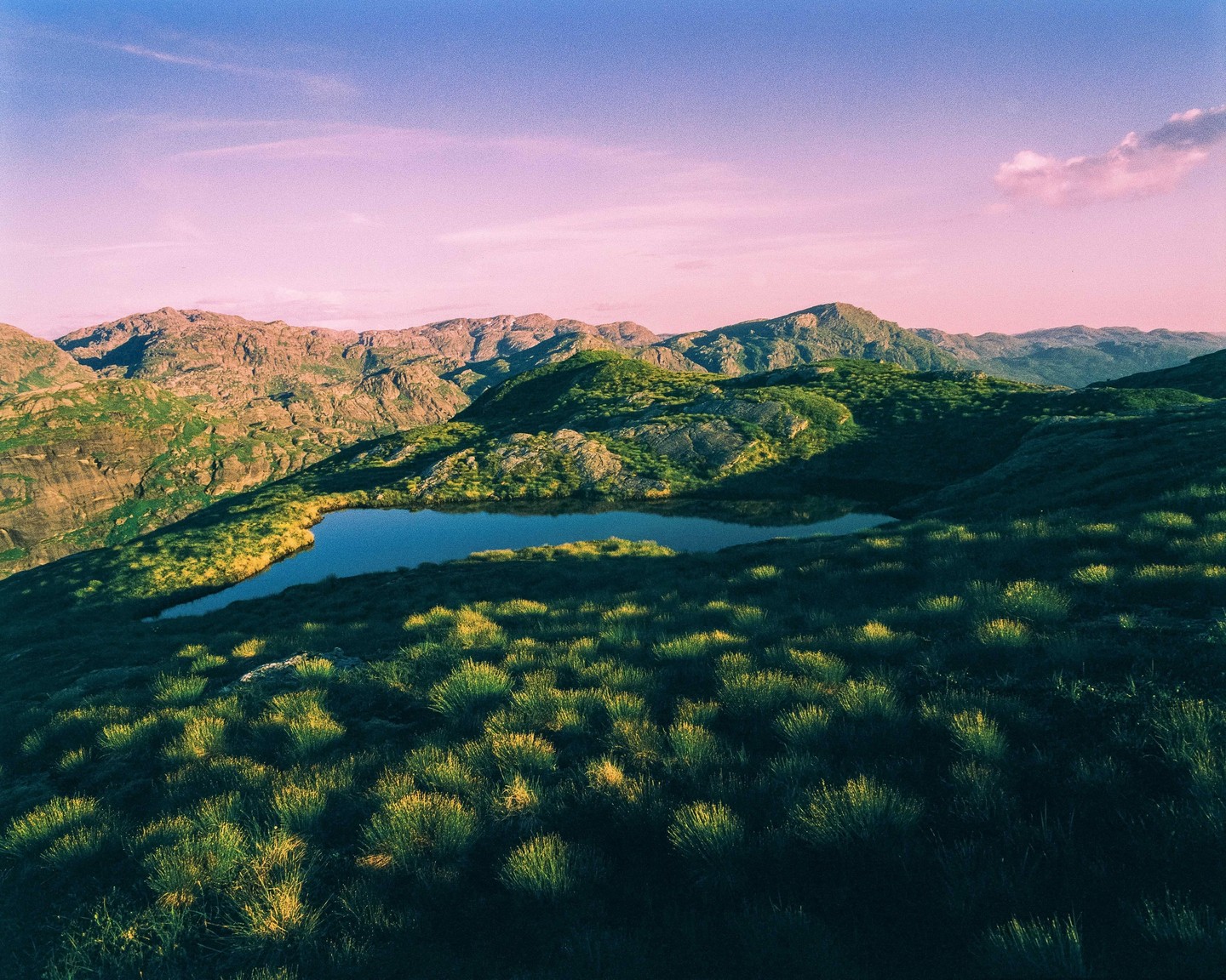 A small lake is nestled among green, rocky mountains under a pink and blue sky.